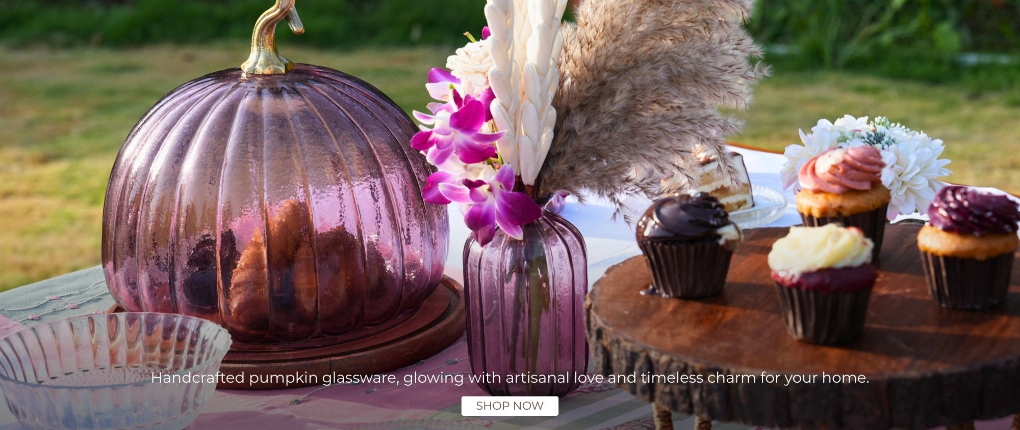 Decorative pumpkin glassware with flowers and cupcakes on a wooden table outdoors.